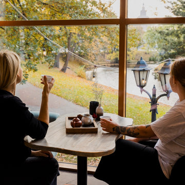 Tejo tējas namiņš Rīgā – cilvēki bauda tēju un sarunas pie kanālmalas loga, piedzīvojot mieru un siltu kopības sajūtu.